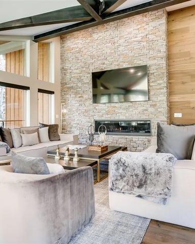 Modern living room with stone fireplace, wall-mounted TV, upholstered seating, wood beams, and large windows overlooking forest.