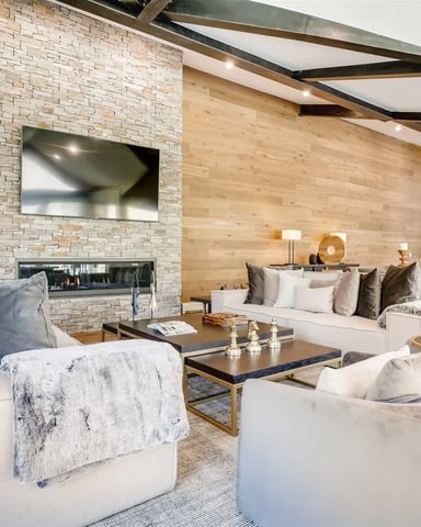 Modern open-plan living room with stone fireplace, TV, cream seating, wood ceiling beams, and skylights