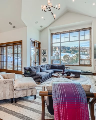 Open-plan living room with vaulted ceiling, stone fireplace, blue sectional sofa, and large windows overlooking water and hillside.