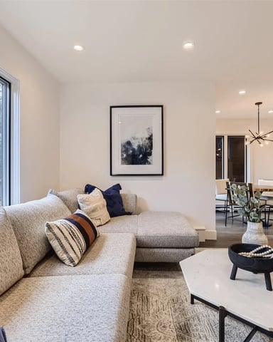 Open-plan living room with beige sectional sofa, large windows overlooking mountains, white coffee table, and kitchen visible in background