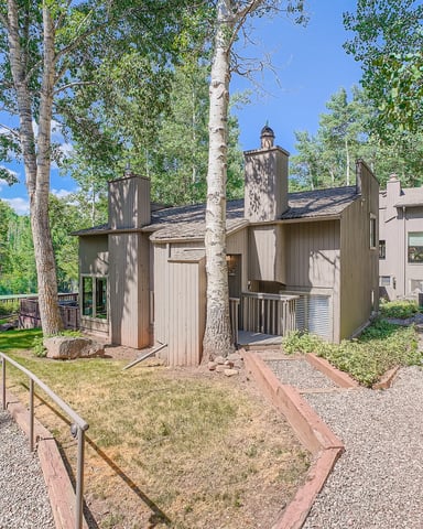 Modern cabin with tall chimneys nestled among aspen and evergreen trees, gravel pathways and metal railings in foreground.