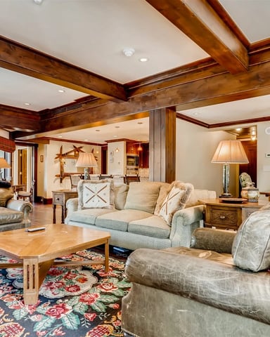 Spacious open-plan living room with exposed wooden beams, multiple seating areas, fireplace, and antler chandelier