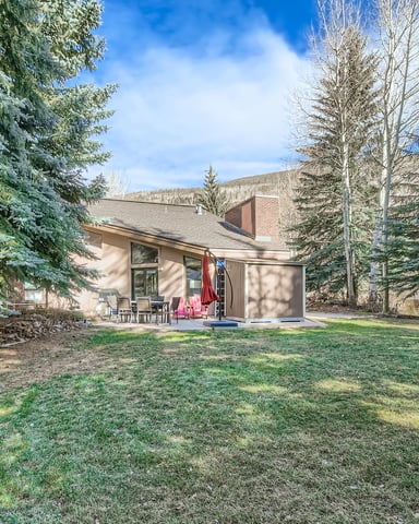 Modern home with brick chimney surrounded by evergreen and aspen trees, patio with umbrella, mountain backdrop.