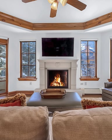 Spacious living room with fireplace, wood beams, multiple windows overlooking forest, and leather seating