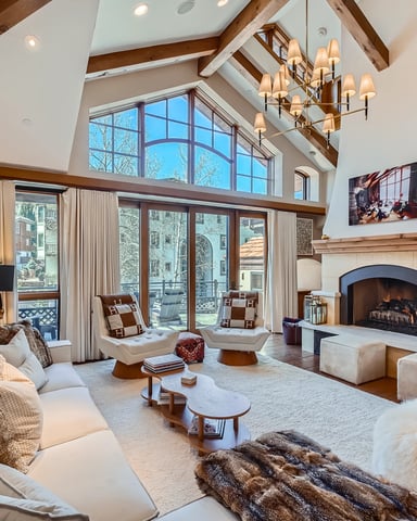 Spacious living room with vaulted wood-beam ceiling, fireplace, large windows, cream seating, and chandelier.