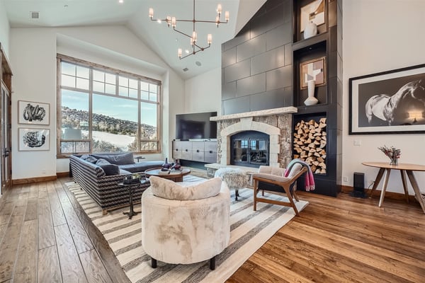 Modern living room with stone fireplace, dark tile accent wall, leather sofa, cream armchairs, wood flooring, and large windows overlooking hillside landscape.