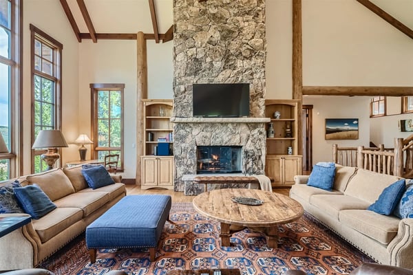 Spacious living room with stone fireplace, exposed wood beams, cream sofas with blue pillows, round wooden table, and large windows overlooking trees.